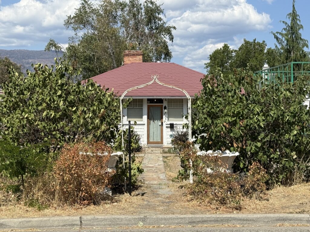 Modest historic cottage at 236 Beach Avenue in Kelowna’s Heritage Conservation Area, with a hipped roof, central entrance, garden and mature landscaping.