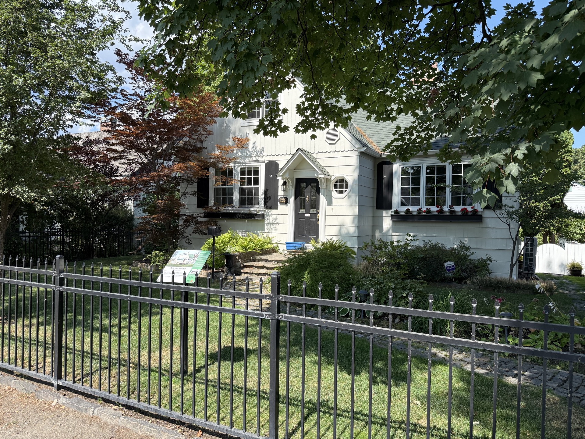White home in Kelowna’s Heritage Conservation Area, shaded by mature trees, with a garden, iron fence and heritage information plaque.